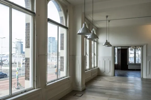 Lichte, lege binnenruimte aan de De Ruijterkade met hoge ramen, hanglampen en uitzicht op moderne kantoorgebouwen.